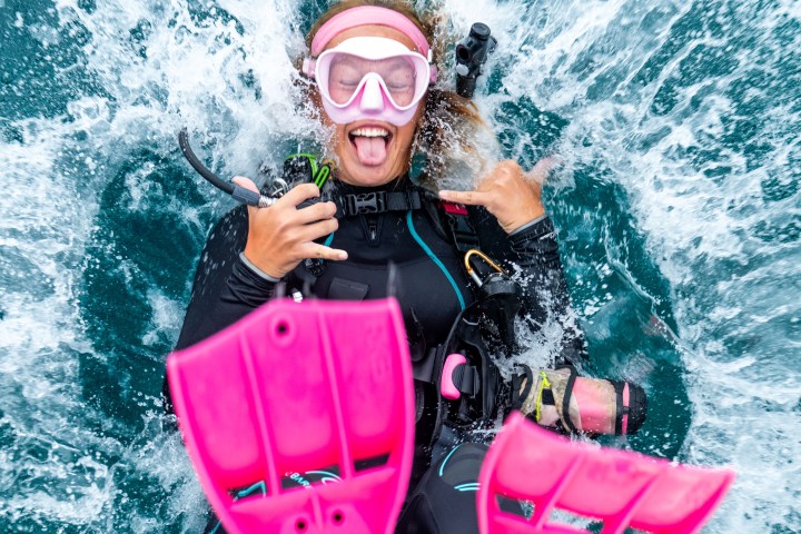 A scuba diver with pink fins and mask splashing into water, making a playful hand gesture.